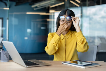 Tired young Indian woman sitting at office desk, holding glasses and rubbing eyes