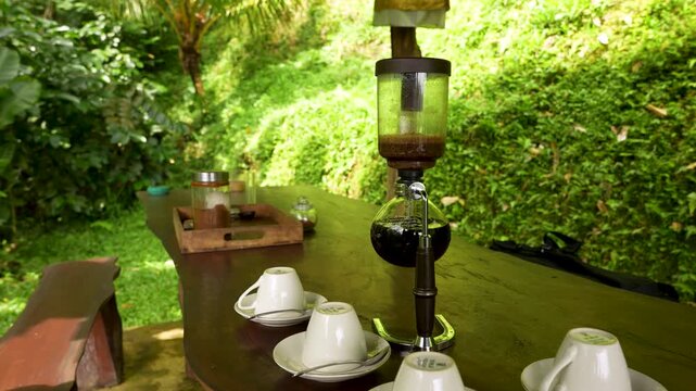 Siphon coffee maker extracts rich kopi luwak beside cups on a rustic table in jungle garden cafe, static handheld shot.