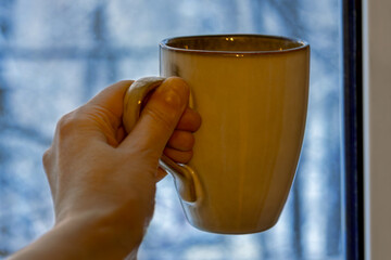 Hand holding warm coffee mug near window on cold winter evening