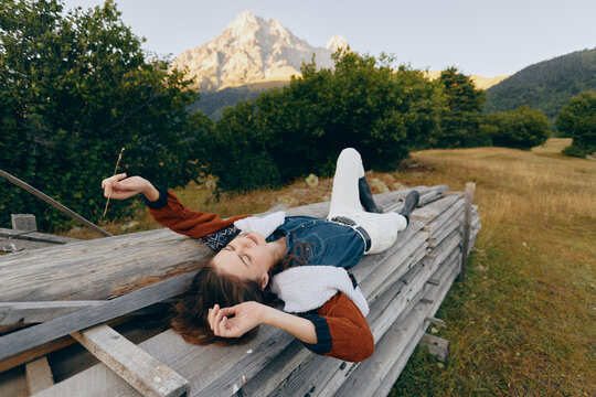 Woman lying relaxed on a wooden fence in a meadow with mountain backdrop and forest, enjoying nature and peaceful outdoor moment in casual outfit during a sunny summer escape.