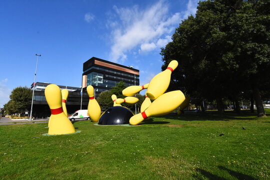 Wide-angle view of the iconic Flying Pins sculpture in Eindhoven, Netherlands. A giant yellow bowling pin and black ball public art installation against a modern building and blue  sky. 