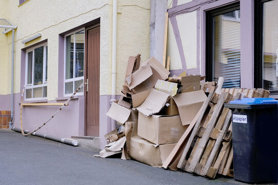 large pile of broken cardboard boxes and wooden pallets against house wall next to paper recycling bin recycling problem, paper waste near building representing environmental contamination and neglect