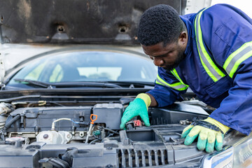 African mechanic wearing blue overalls and green gloves inspecting a car engine under an open hood, diagnosing a mechanical issue.