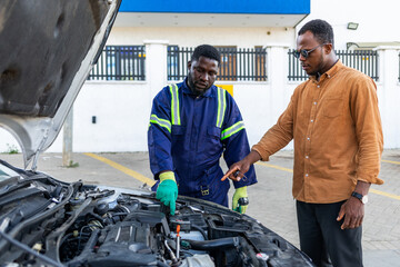 African mechanic in blue overalls explains an engine issue to a customer in his workshop