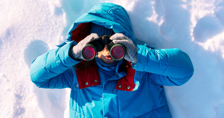 young woman wearing a blue winter jacket and gloves with surprised expression looks through binoculars while lying in snow. The scene captures a winter exploration theme