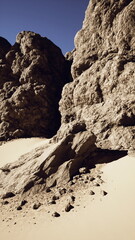 Majestic rocky formations tower over a soft sandy area, creating a striking contrast. The bright skies reflect a serene moment in the desert, showcasing natures beauty and strength.