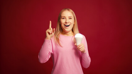 Cheerful smiling woman holding lamp pointing finger up having good idea isolated on red background