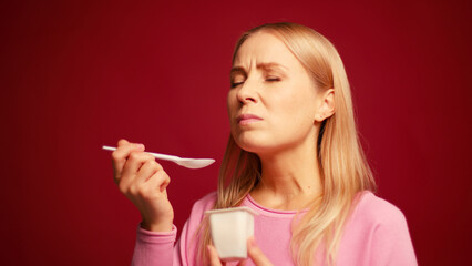 Obraz premium Smiling blonde woman holding white cup with yogurt eating tasty dessert enjoying on red background