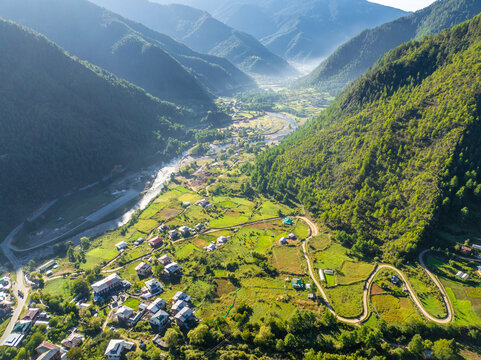 Aerial view of a winding road snakes through verdant hillsides towards a valley dotted with small buildings and a flowing river, haa valley, Paro, Bhutan.