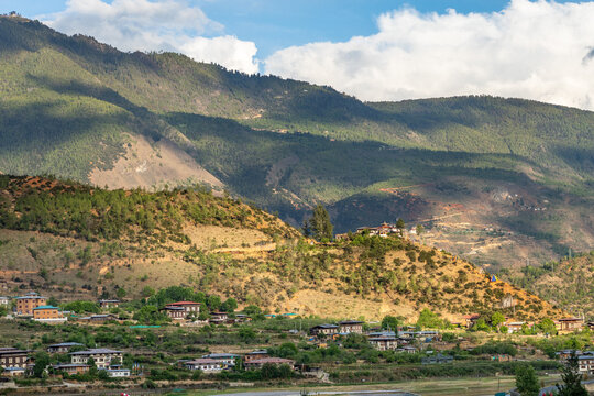 View of the majestic mountains cascading down to meet a cluster of buildings nestled in the valley under a sky of fluffy clouds, Paro, Paro, Bhutan.