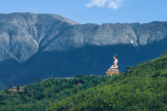 View of golden Buddha statue perched atop a vibrant green hill, contrasting against a backdrop of majestic, shadowed mountains, Thimphu, Thimphu, Bhutan.