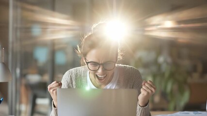 A woman at her desk expresses excitement with raised fists, symbolizing a moment of achievement. The focused atmosphere is enhanced by a laptop and warm lighting behind her.