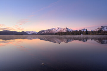Calm winter reflections in Derwentwater lake at sunrise with snow on mountains, Lake District, England, UK.