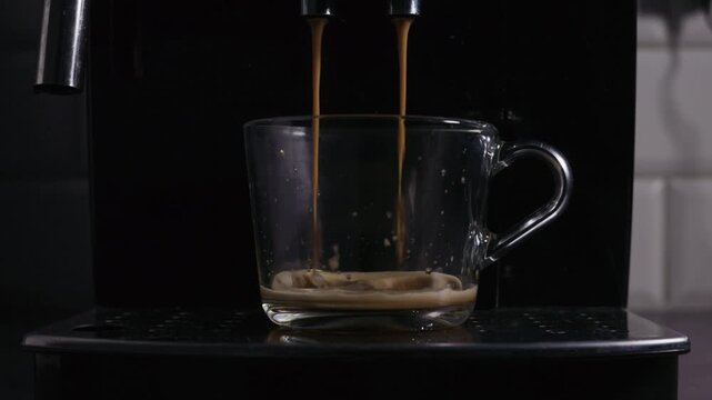 A close-up view of a glass cup being filled with freshly brewed espresso from a coffee machine. The rich crema forms on top as the dual streams of coffee pour smoothly into the transparent mug.