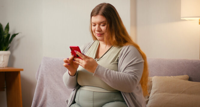 Plus size woman scrolls smartphone while sitting on couch