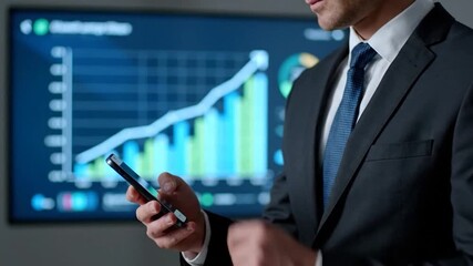 A man in a suit looks at his phone while a financial graph rises on a monitor - Powered by Adobe