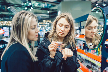 Two cheerful friends are choosing mascara and lipstick in cosmetics store. High quality photo