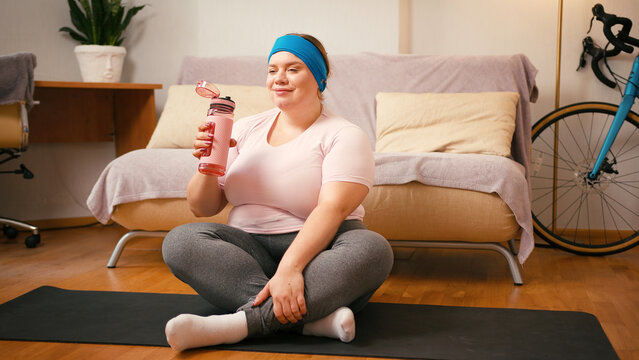 Plus size woman drinks water on yoga mat during workout break - Powered by Adobe