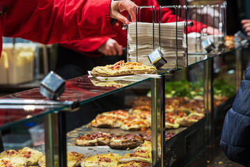 Vendor in a red jacket serving a pizza snack at a glass counter with hot dishes at winter market, grabbing a napkin from a stack nearby preaparing a meal for customers
