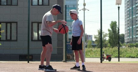 Father teaches son basketball on outdoor court. Family training creates positive sport mood. Child boy learns basketball skills in summer. Father supports learning process. builds teamwork. - Powered by Adobe
