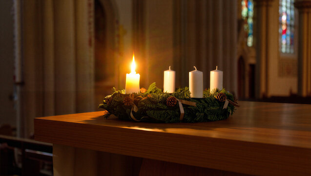 A dimly lit advent wreath with three unlit white candles and one lit yellow candle sits on a wooden surface in a church setting First Sunday of Advent