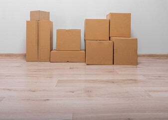 A collection of stacked, plain brown cardboard moving or shipping boxes rests on a light wood laminate floor against a white or light gray wall.