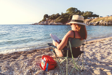 Woman in sunhat reads book on a folding chair by the ocean, enjoying peaceful summer beach vibes.