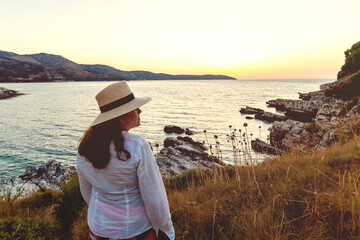 Rear view of a woman in a sunhat admiring the sea at sunset, standing on rocky shore.
