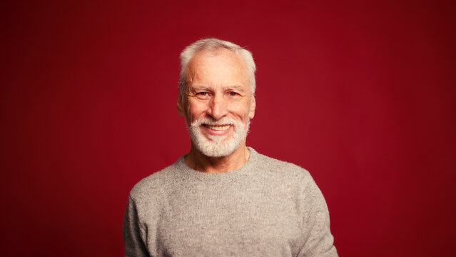 Portrait of happy senior man touching grey beard looking at camera posing isolated on red background
