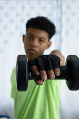 Brown skinned teenager in close up performing a front dumbbell raise, wearing gloves and training in the living room of his home. Concept of exercise and at home workout