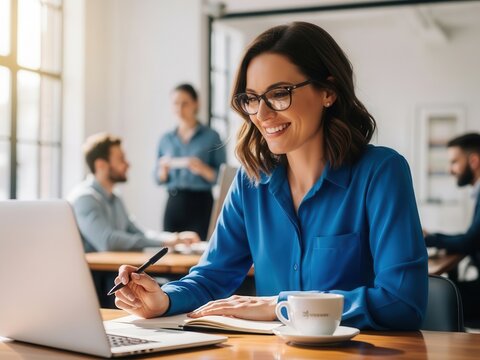 Woman in blue shirt working on laptop and writing in notebook