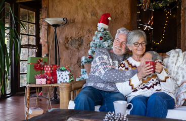 Joyful older couple in festive sweaters enjoying a cozy Christmas moment together sitting on sofa with holiday lights, wrapped gifts, and Christmas tree. radiating love, comfort and emotions