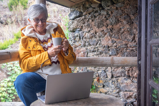 Cheerful senior woman works on her laptop while enjoying a warm drink on a rustic outdoor terrace. Dressed in a cozy sweater and bright yellow jacket, she relaxes in a natural mountain setting - Powered by Adobe