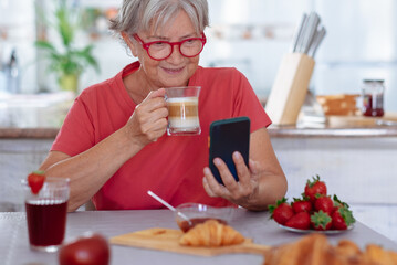 Smiling relaxed senior woman while drinking a coffee with milk sitting at home using phone. Elderly grandmother enjoying breakfast with fruit and croissant in home kitchen