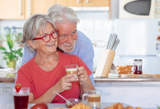 Cheerful affectionate senior Caucasian couple hugging and smiling while having breakfast at home. Relaxed and happy seniors enjoying healthy nutrition