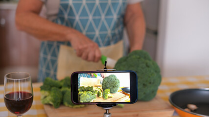 Elderly woman wearing apron chops fresh broccoli on a kitchen counter recording a video by smartphone. The scene highlights healthy meal preparation, emphasizing fresh vegetables, home cooking