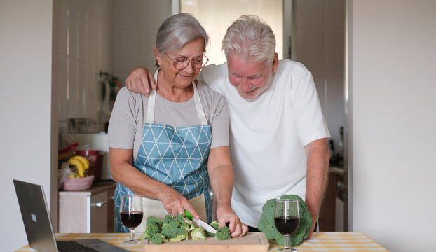 Elderly happy couple on a kitchen counter chops fresh broccoli while enjoying a red wineglass. Concept of healthy meal preparation, fresh vegetables, home cooking together - Powered by Adobe