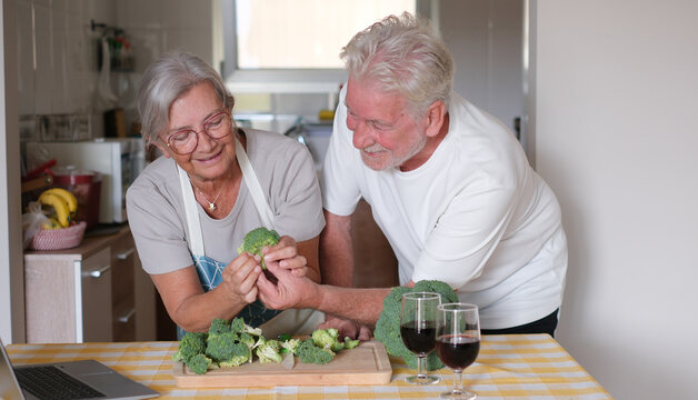 Elderly happy couple on a kitchen counter chops fresh broccoli while enjoying a red wineglass. Concept of healthy meal preparation, fresh vegetables, home cooking together