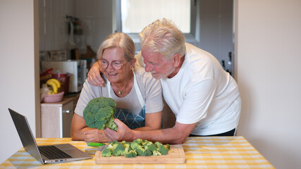 Happy older couple at the kitchen counter chopping fresh broccoli while drinking a glass of red wine and looking recipes on their laptop. Concept of preparing healthy meals and home cooking together