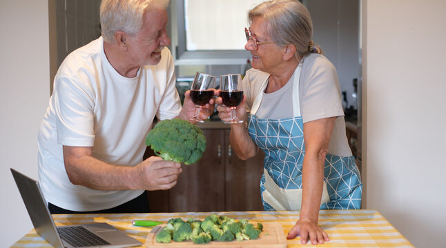 Elderly happy couple on a kitchen counter chops fresh broccoli while enjoying a red wineglass. Concept of healthy meal preparation, fresh vegetables, home cooking together - Powered by Adobe