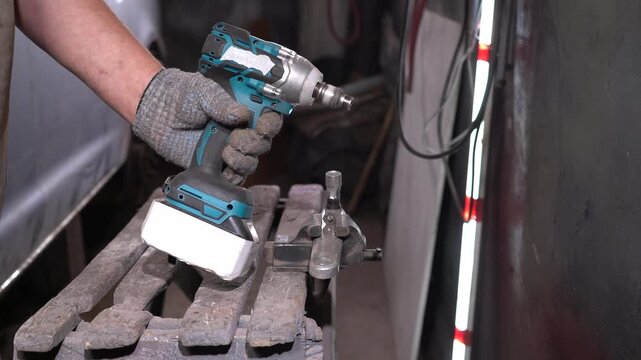 A person uses a cordless impact wrench to perform metal assembly work in a workshop filled with tools and materials during daylight. The workspace is well-lit.