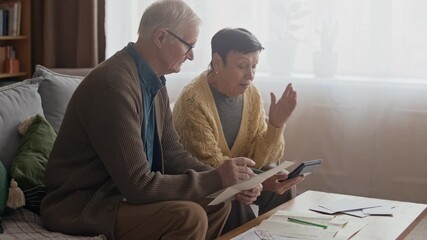 Medium shot of elderly Caucasian couple calculating household expenses and bills on sofa using calculator, dealing with financial difficulties in cozy living room at home - Powered by Adobe