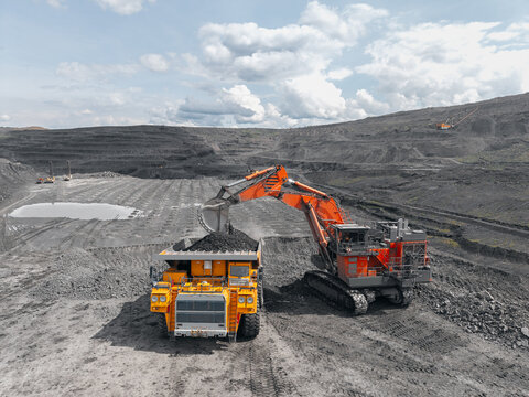 Industrial excavator loads coal into back of yellow heavy dump truck in open pit mine under bright sky aerial view, concept mining industry