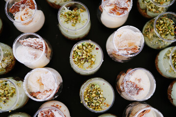 Overhead view of numerous individual dessert shots in small glass cups, featuring layered cream and pistachio garnishes, arranged randomly on a dark surface