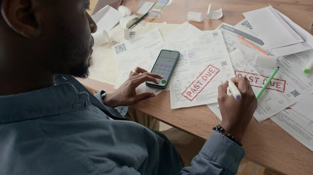 Over the shoulder of Black man reviewing and organizing household expenses while using calculator on smartphone, focusing on budgeting and managing bills at home