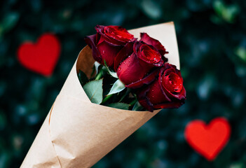Bouquet of deep red roses wrapped in craft paper, accented by heart shapes in lush green background