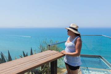 Woman on balcony overlooking ocean with boat trail under clear blue sky in tropical setting.