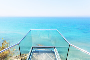 Transparent glass deck extending over cliff with panoramic ocean view and turquoise water under sunny sky.