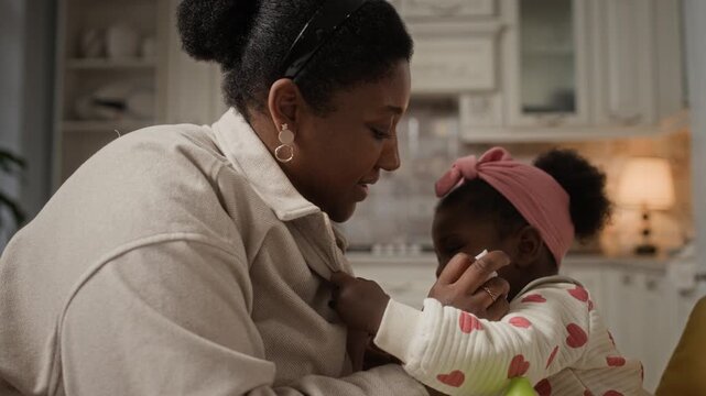 Loving Black mother calming her crying baby, wiping her face with napkin and hugging her while sitting in kitchen