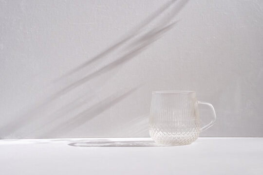 Transparent textured glass mug on a white surface with soft natural sunlight casting long shadows on a light wall creating a minimal calm modern aesthetic scene. Monochrome